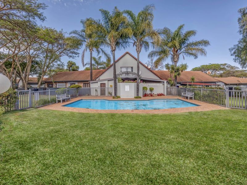 A well-maintained outdoor pool area featuring a paved surround, perimeter fencing, and established palm trees set within a landscaped garden.
