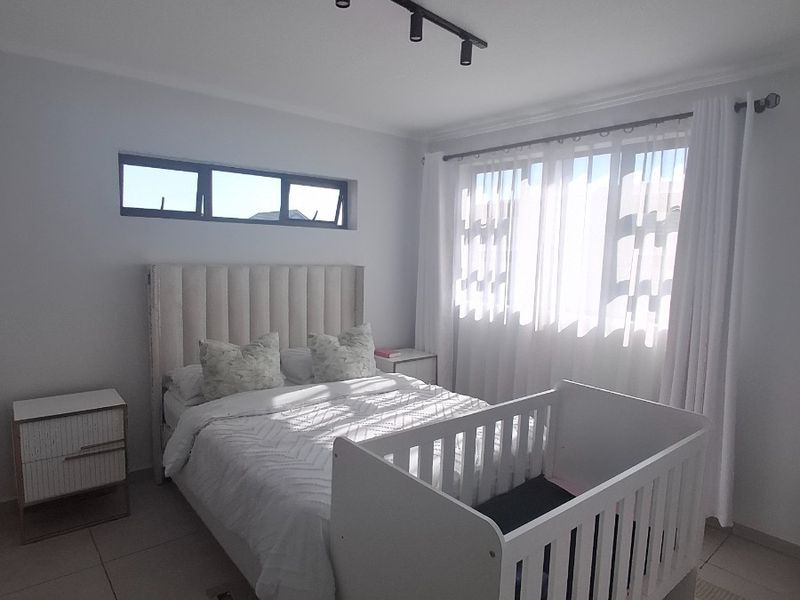 A bright, modern bedroom featuring neutral tiled flooring, contemporary track lighting, and a combination of large windows and a clerestory window for natural light.
