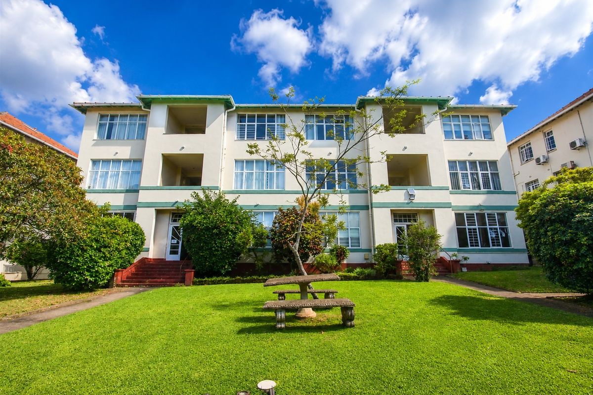 A view of the rear facade of a multi-story apartment building featuring a well-maintained communal garden and paved walkways.
