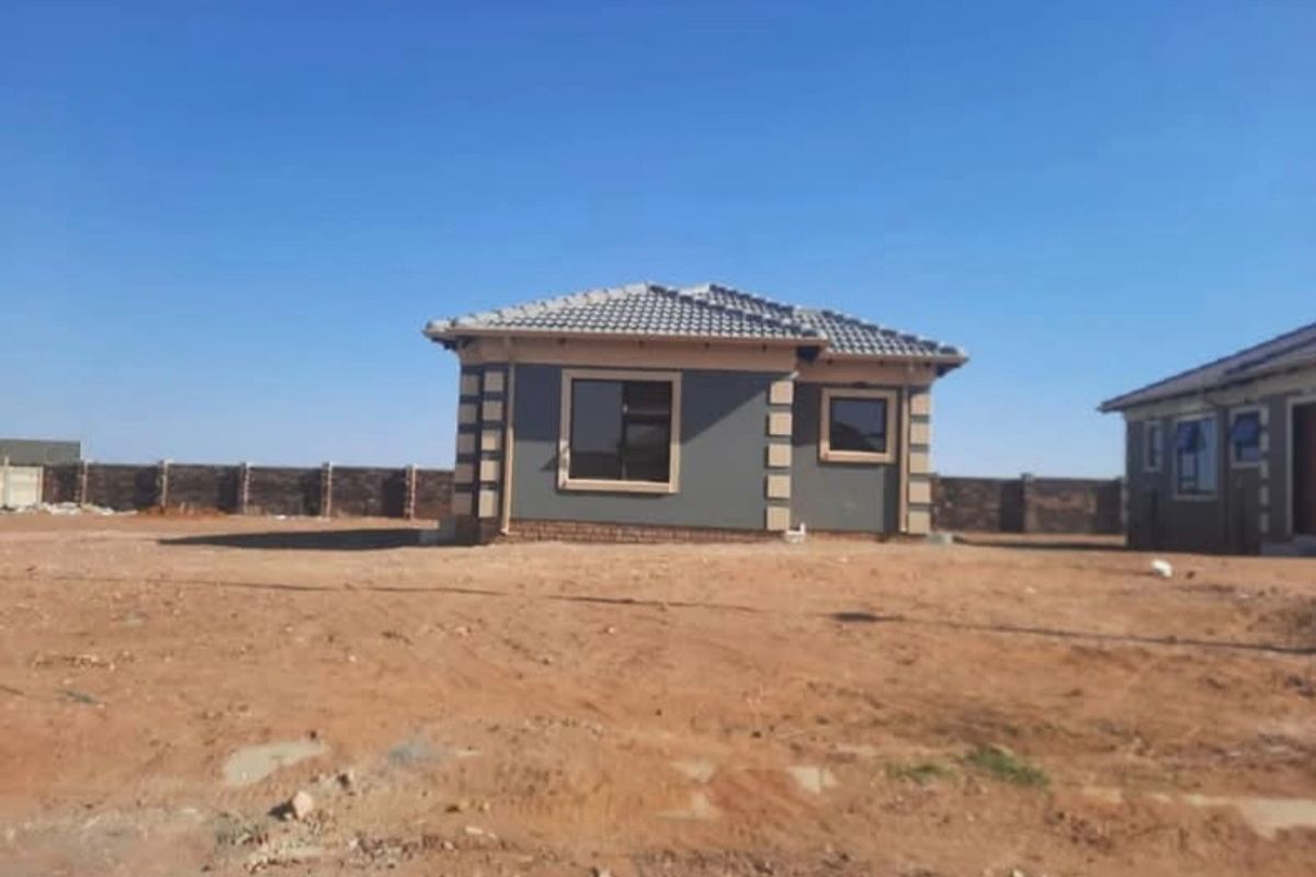 A newly constructed single-story house featuring a tiled roof and modern grey facade, situated on an unlandscaped plot with perimeter walling.