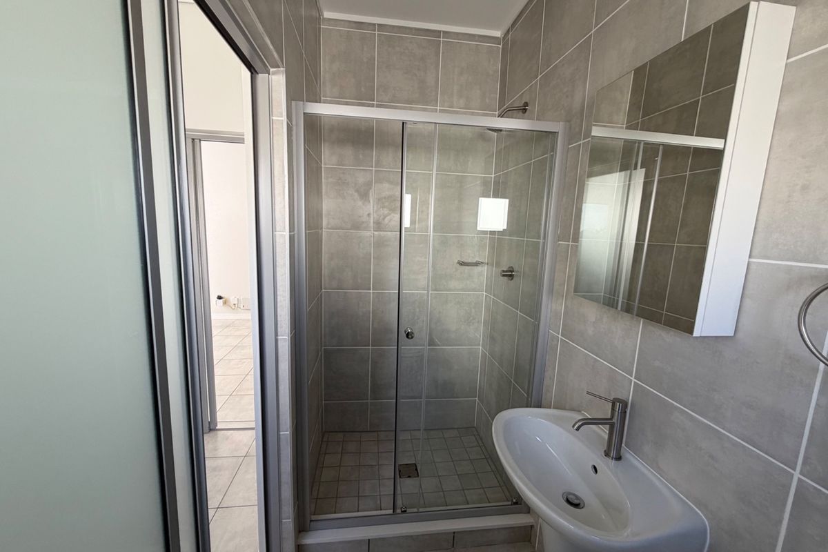 A clean, modern bathroom featuring a glass-enclosed shower, pedestal basin, and wall-mounted mirror cabinet with neutral grey wall and floor tiling.