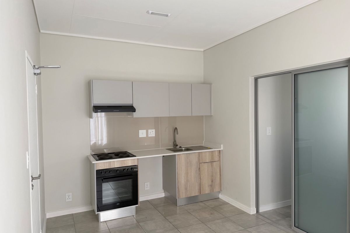 A modern, compact kitchen featuring neutral cabinetry, built-in oven and hob, stainless steel sink, and tiled flooring, presented in a clean, empty space.