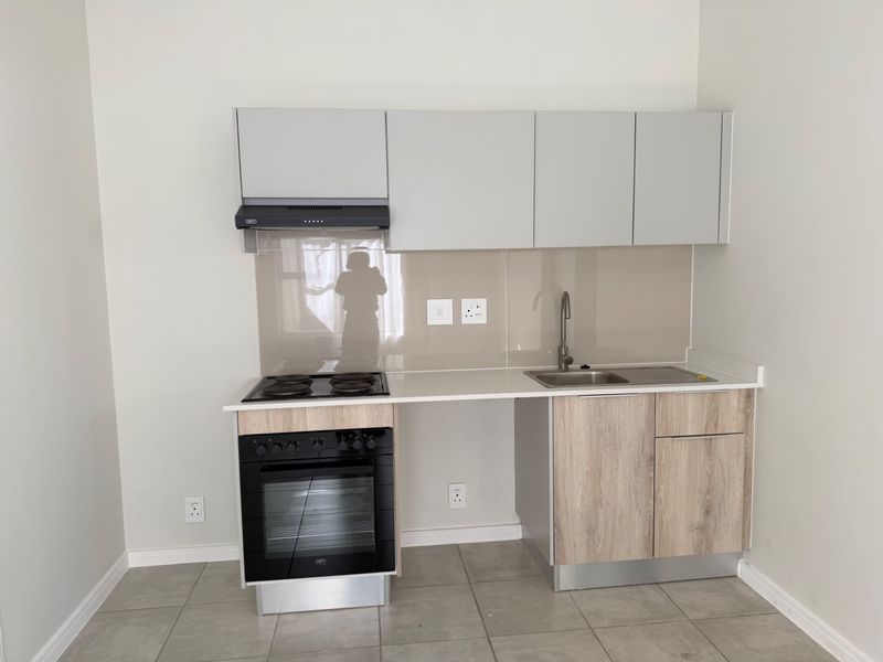 Modern compact kitchen featuring light wood-finish cabinetry, stone countertops, a built-in oven with hob, and a sleek glass splashback.