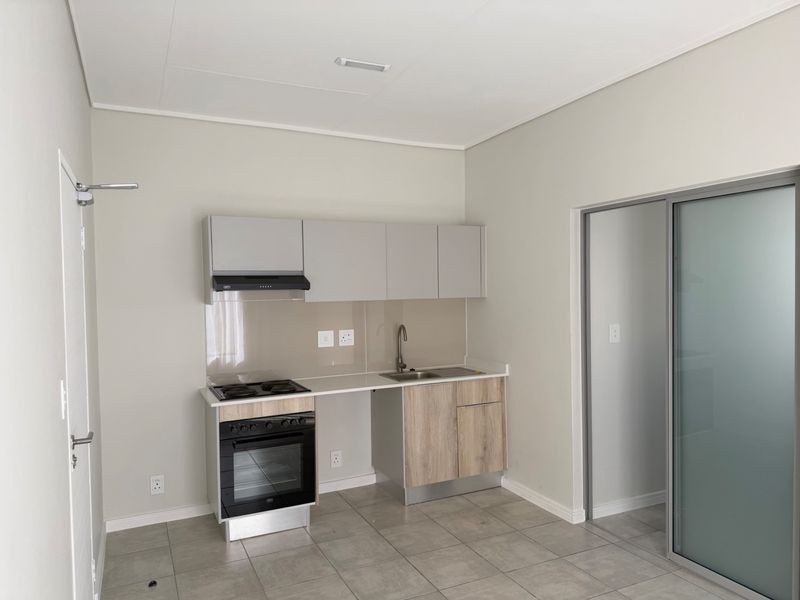 A modern, compact kitchen featuring neutral cabinetry, built-in oven and hob, stainless steel sink, and tiled flooring, presented in a clean, empty space.
