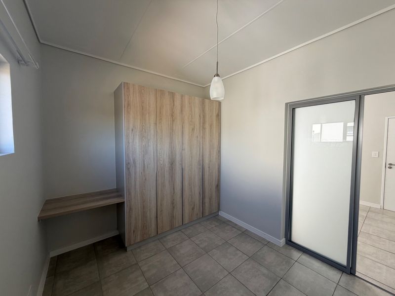 A clean, modern bedroom featuring built-in wood-finish cupboards, an integrated desk, tiled flooring, and a frosted glass door.