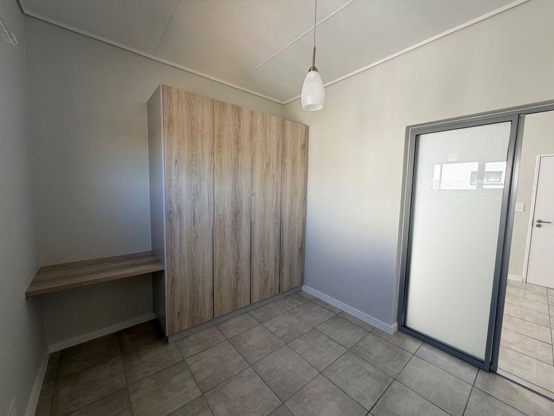 A clean, modern bedroom featuring neutral tiled flooring, built-in wood-finish cupboards, an integrated desk, and a stylish pendant light fixture.