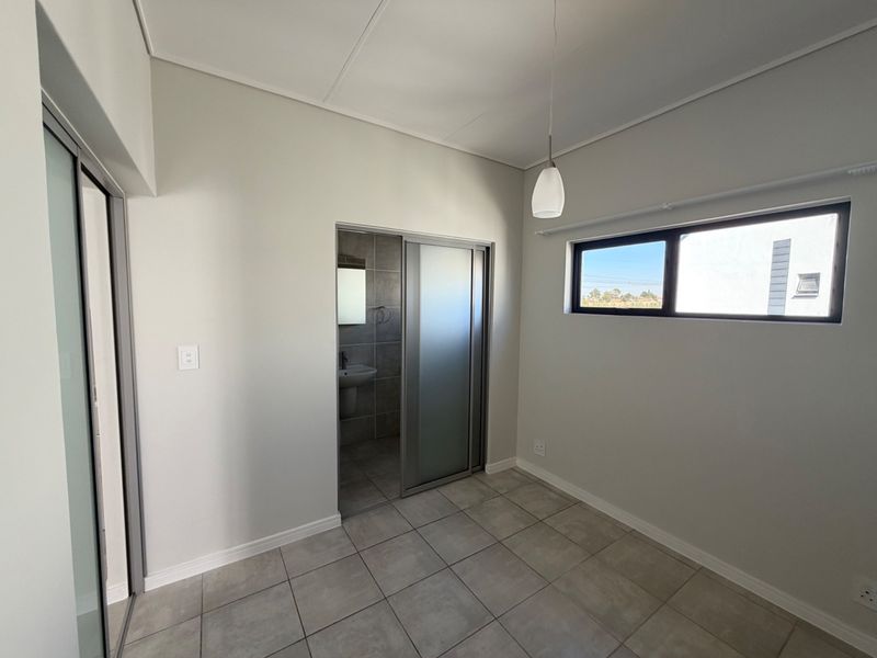 A clean, modern bedroom featuring neutral tiled flooring, a sliding door leading to an en-suite bathroom, and a contemporary pendant light fixture.