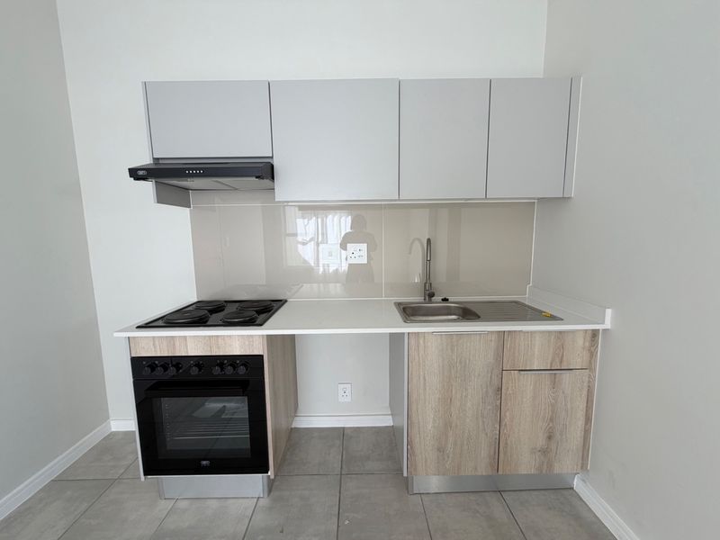 A modern, compact kitchen featuring light wood-finish cabinetry, stone countertops, a built-in oven with electric hob, and a sleek glass splashback.