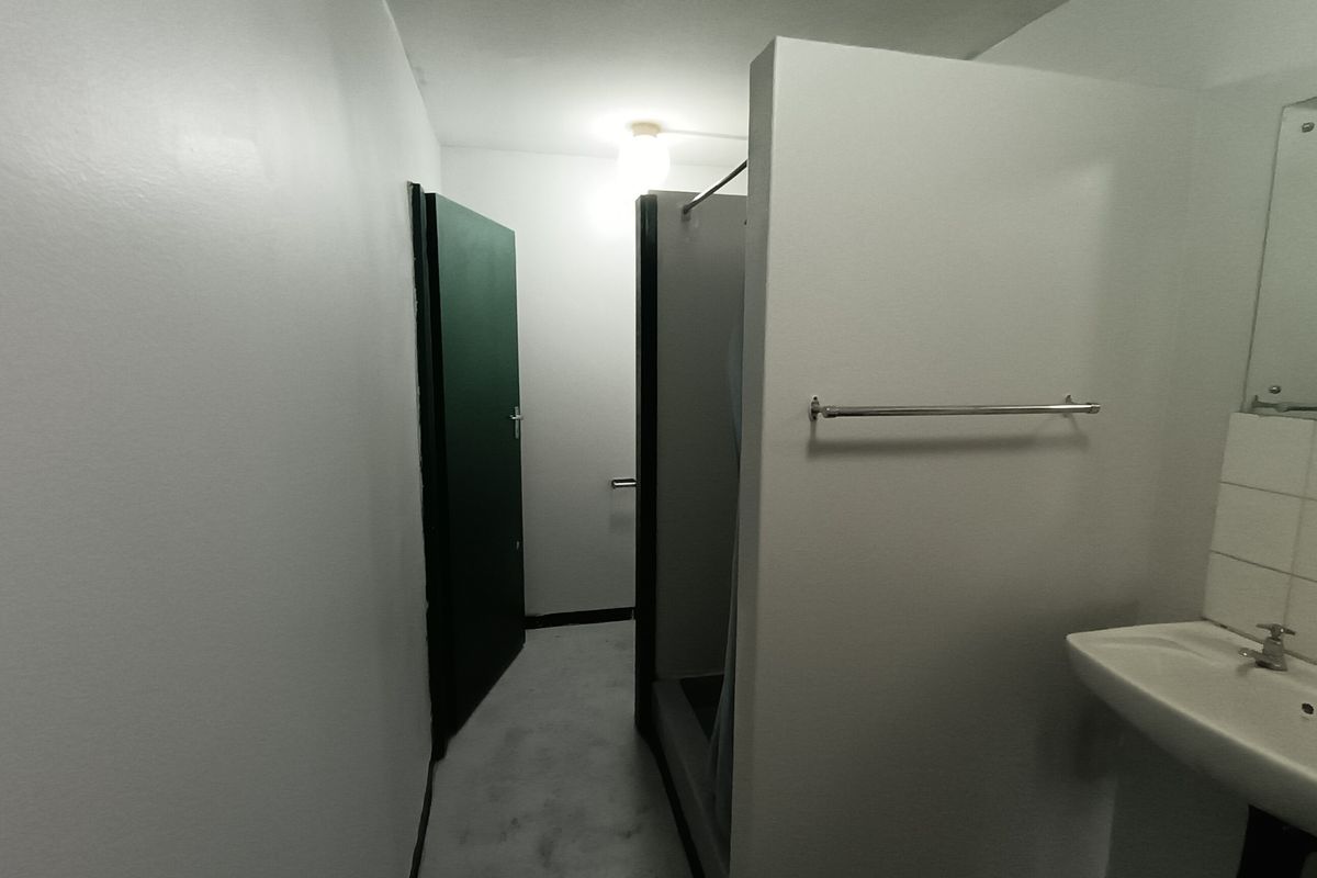 A functional industrial-style bathroom featuring a wall-mounted basin, shower partition, and basic fixtures, presenting a clean but dated aesthetic.
