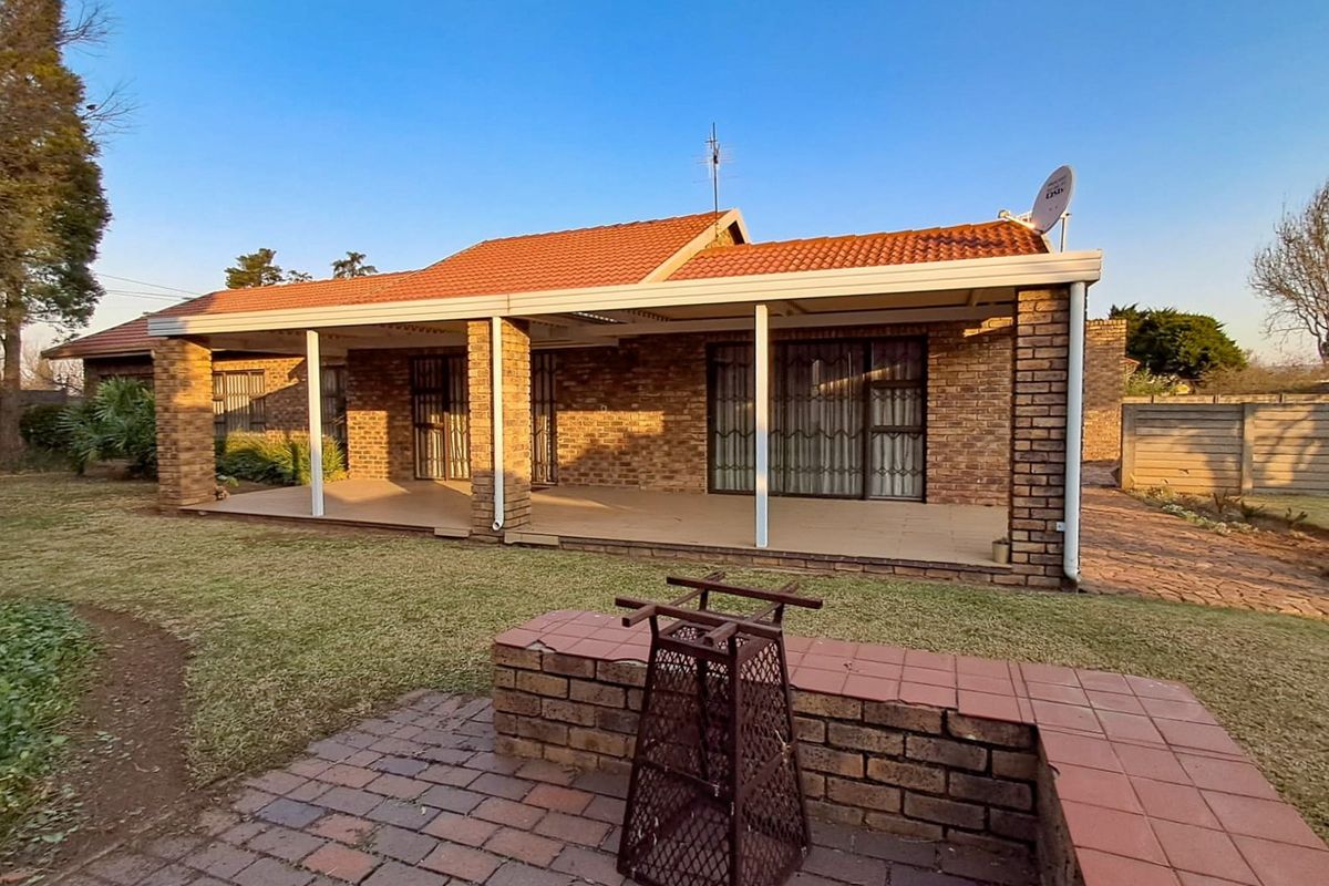 A well-maintained rear exterior view featuring a spacious covered patio, brick facade, tiled roof, and a built-in brick braai area in the garden.