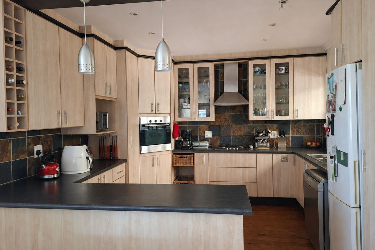A functional kitchen featuring light wood cabinetry, dark laminate countertops, a slate tile backsplash, and integrated appliances including an oven and extractor fan.