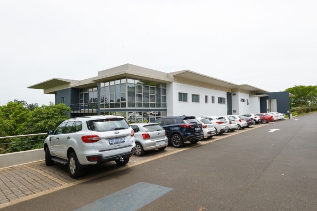 Modern industrial office building featuring a large glass facade, concrete roof overhang, and extensive paved parking area.
