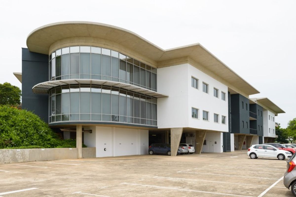 Modern industrial office building featuring a curved glass facade, concrete cantilevered roof, and ground-level parking area.