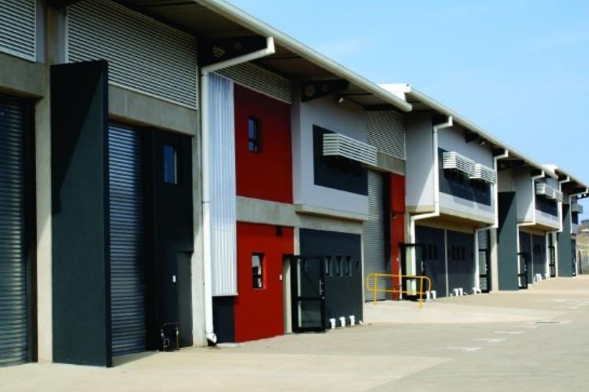 Modern industrial warehouse complex featuring multiple units with roller shutter doors, concrete apron, and contemporary red and grey facade finishes.