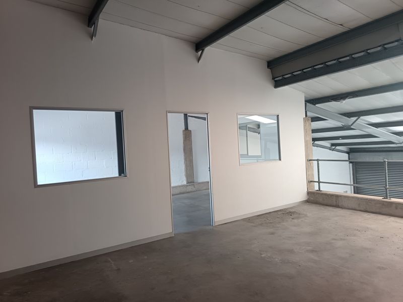 An empty industrial office space featuring exposed steel beams, concrete flooring, and internal windows overlooking the warehouse area.