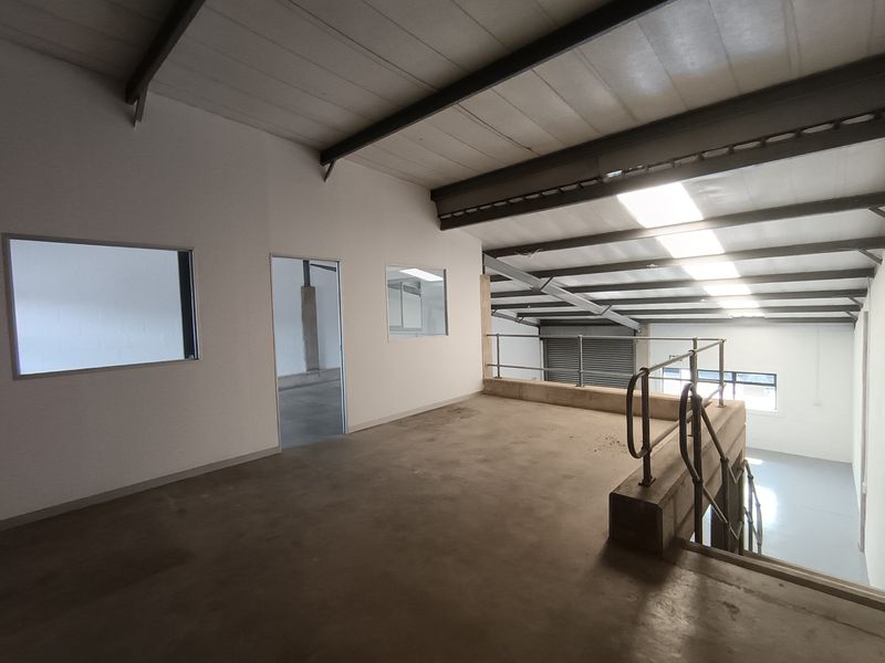 Spacious industrial mezzanine level featuring exposed steel roof trusses, skylights, concrete flooring, and a metal staircase with safety railings.