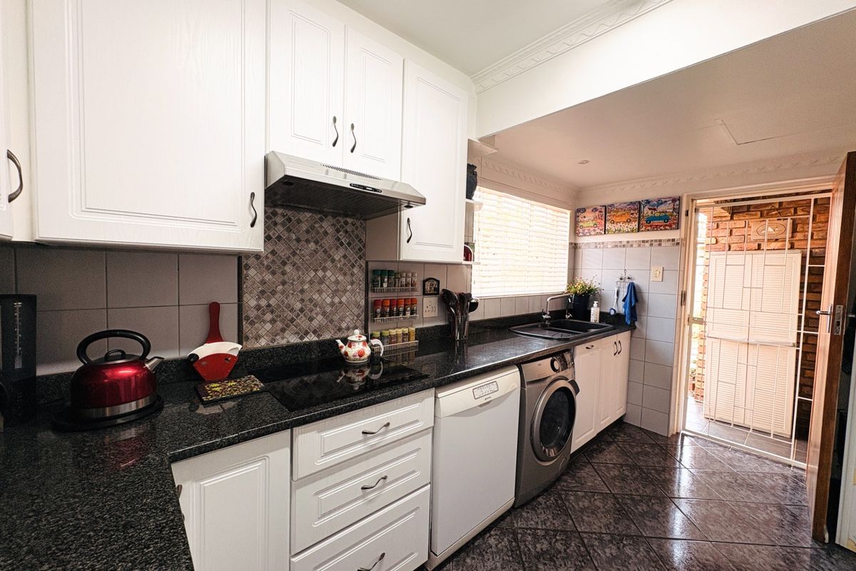 A functional kitchen featuring white cabinetry, granite countertops, an electric hob with extractor fan, and integrated space for laundry appliances, leading to an exterior area.