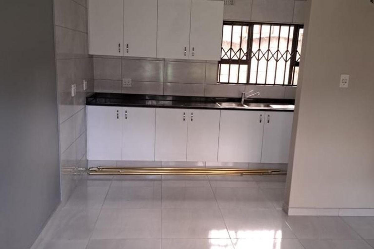 A clean, empty kitchen featuring white built-in cabinetry, dark granite countertops, a stainless steel double sink, and neutral tiled flooring.