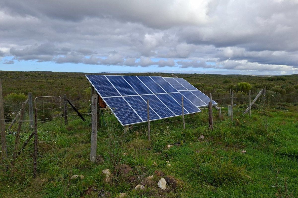 A ground-mounted solar panel array situated in a rural field, enclosed by wire fencing and wooden posts.