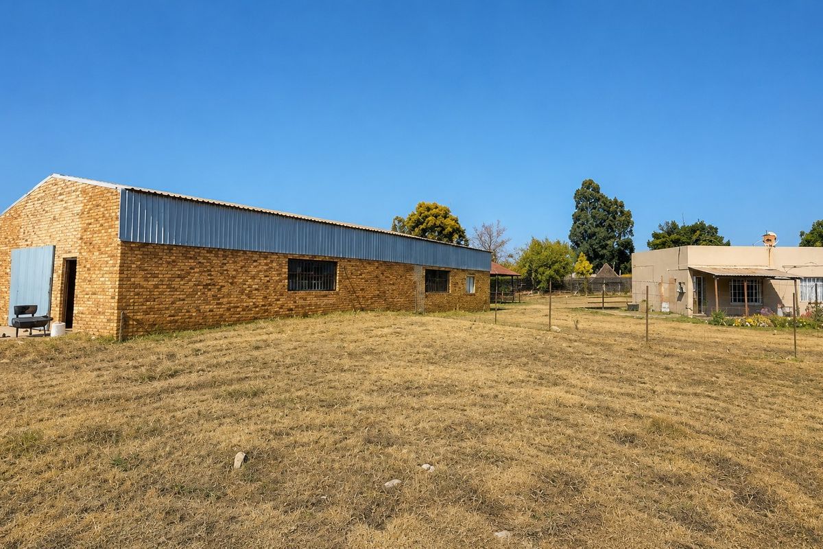 View of the property grounds featuring a large face-brick outbuilding and a separate single-story dwelling set within a spacious, dry garden area.