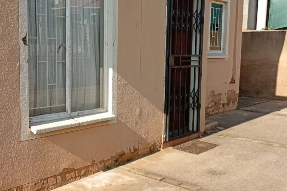 Rear exterior view of the property featuring concrete paving, a security gate, and visible wear to the exterior wall plaster and paint finish.