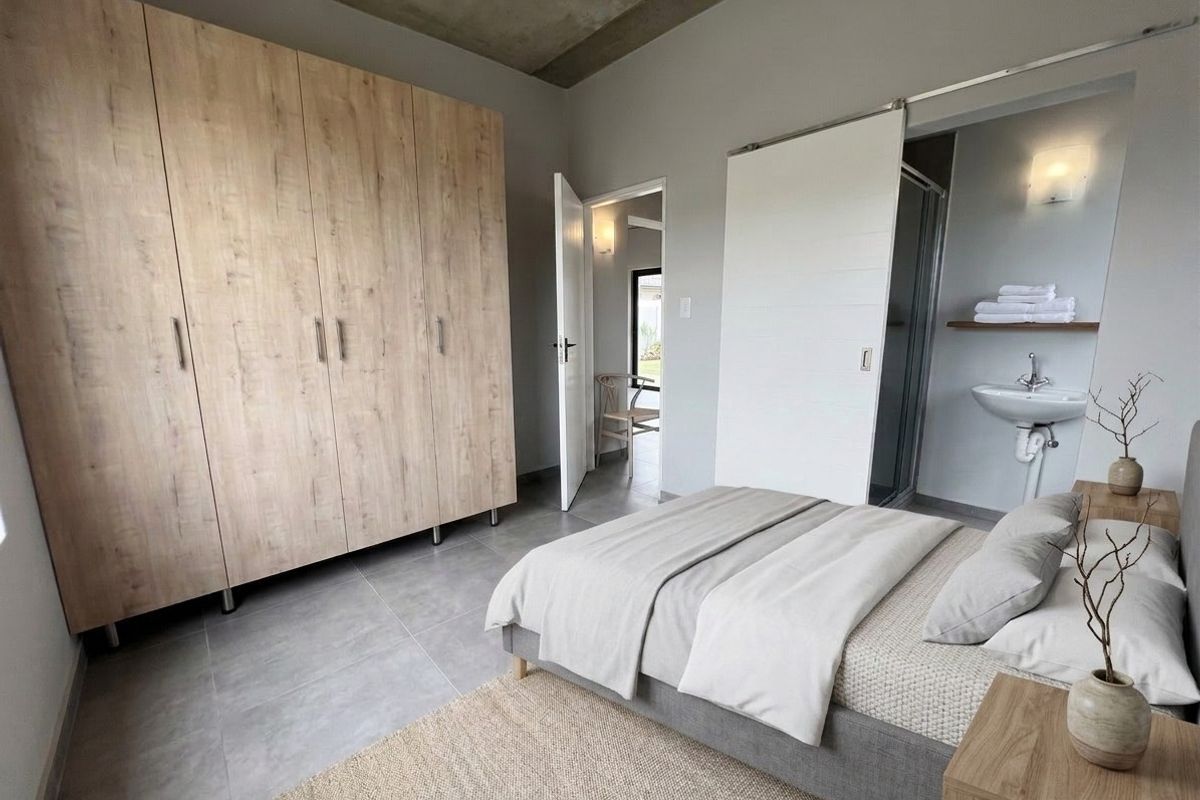 Modern bedroom featuring built-in wood-finish wardrobes, exposed concrete ceiling, tiled floors, and an en-suite bathroom with a wall-mounted basin.
