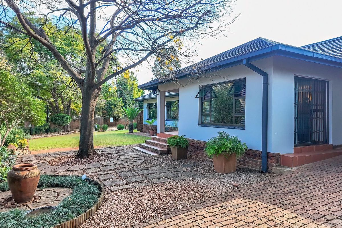 A well-maintained rear exterior view featuring a paved patio area, mature trees, and a landscaped garden with brick and tile finishes.
