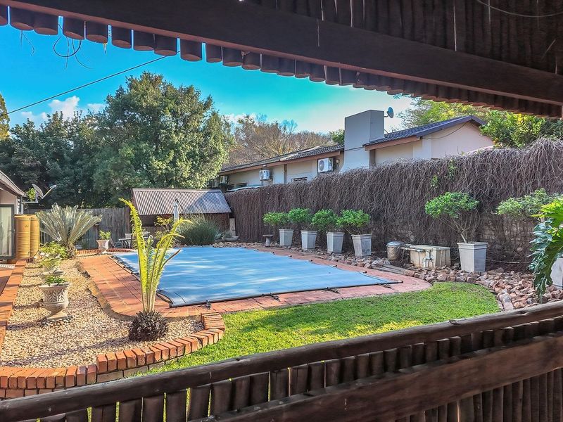A view of a backyard swimming pool with a cover, surrounded by brick paving and a landscaped garden, viewed from a covered patio area.