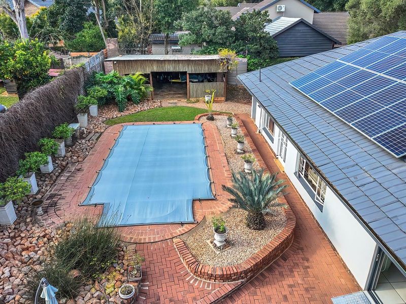 Aerial view of a residential backyard featuring a swimming pool with a cover, brick paved patio, and solar panels installed on the tiled roof.