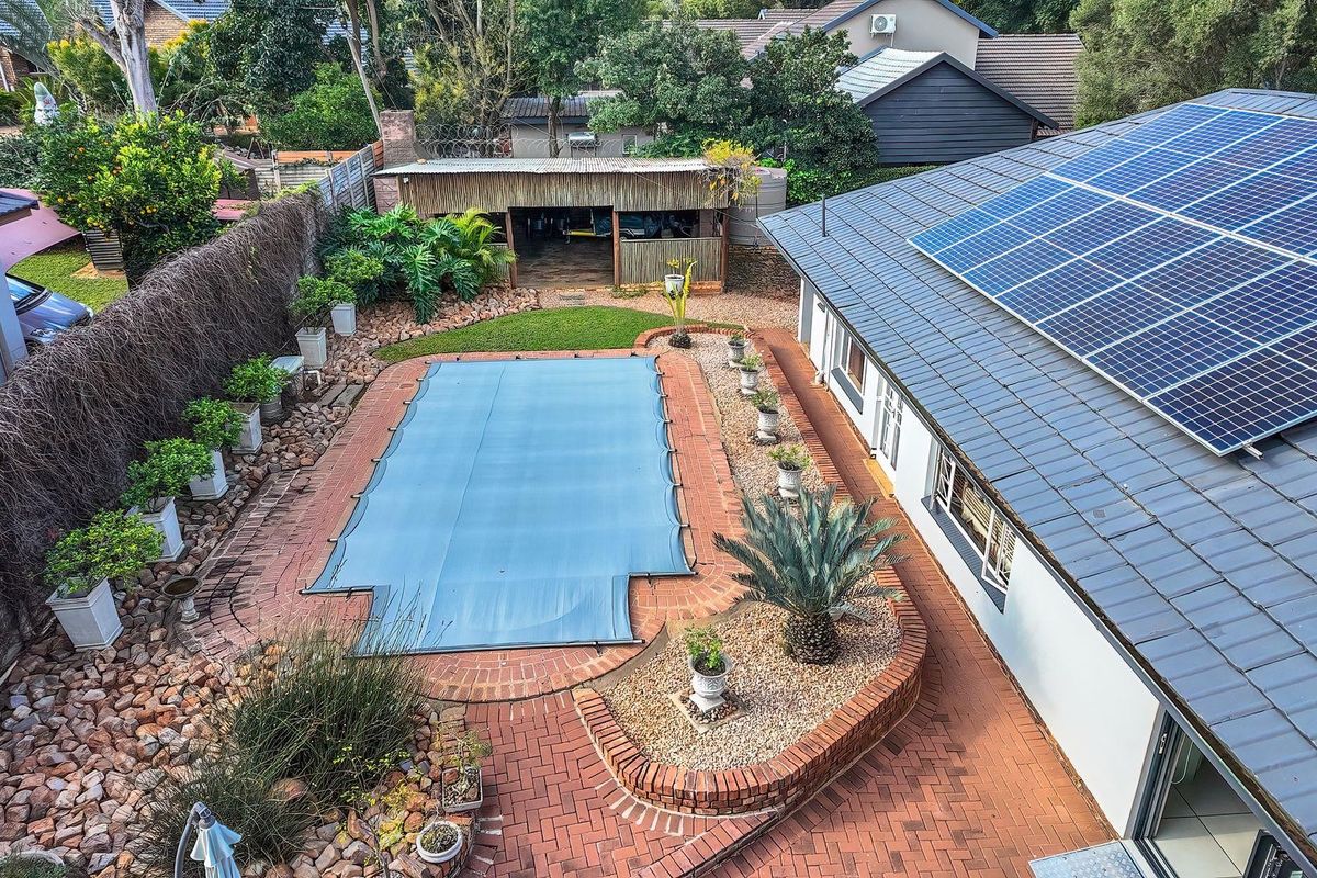 Aerial view of a residential backyard featuring a swimming pool with a cover, brick paved patio, and solar panels installed on the tiled roof.