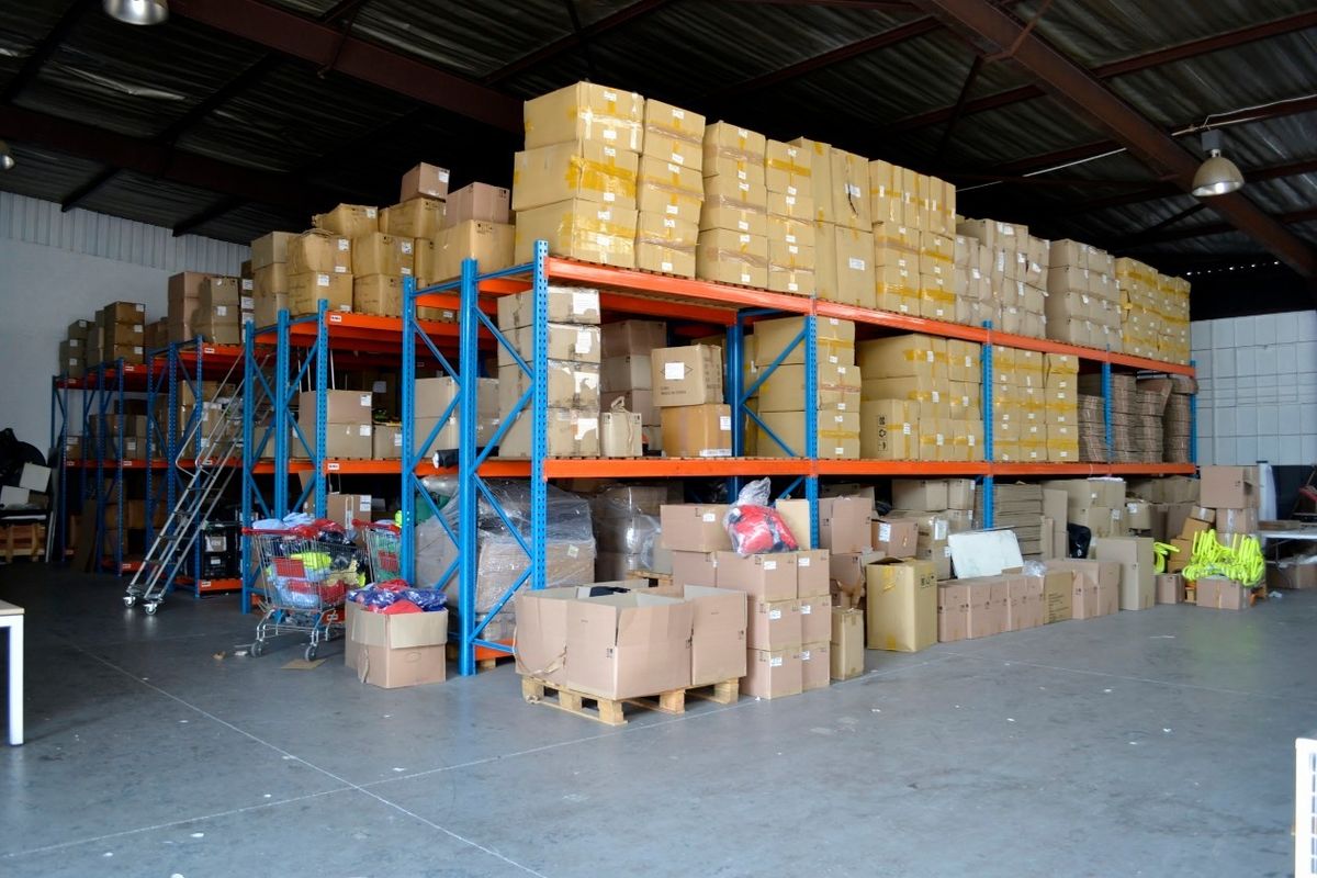 Spacious industrial warehouse featuring high ceilings with exposed steel trusses, concrete flooring, and extensive heavy-duty steel racking systems for storage.