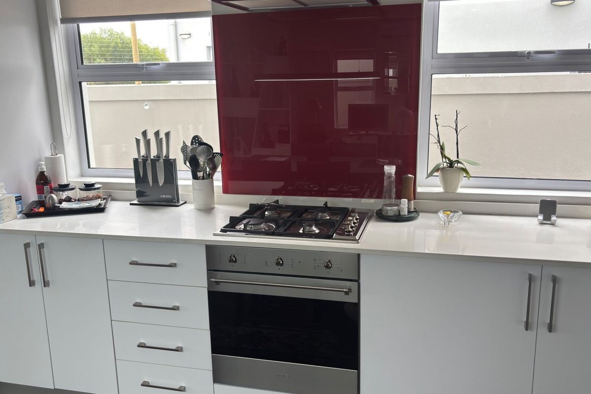 Modern kitchen featuring white cabinetry, stone countertops, a gas cooktop with a red glass splashback, and large windows providing natural light.