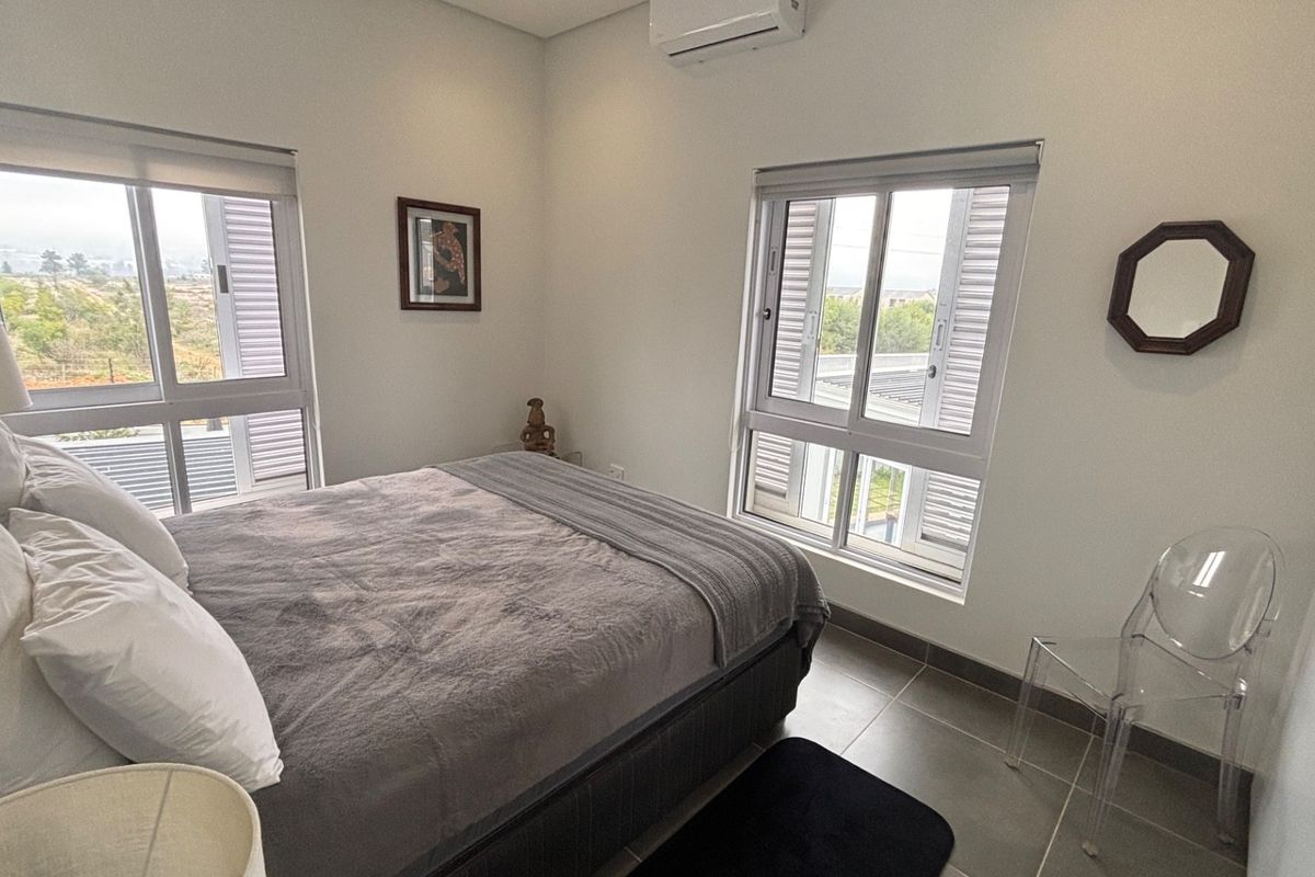 A bright, modern bedroom featuring tiled floors, split-system air conditioning, and large windows with roller blinds, offering a clean and functional living space.