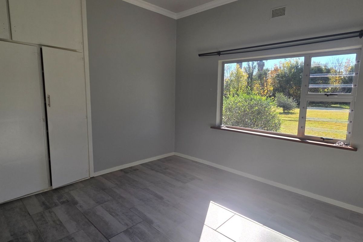 A bright, empty bedroom featuring neutral grey walls, modern laminate flooring, and built-in storage cupboards with a view of the garden.