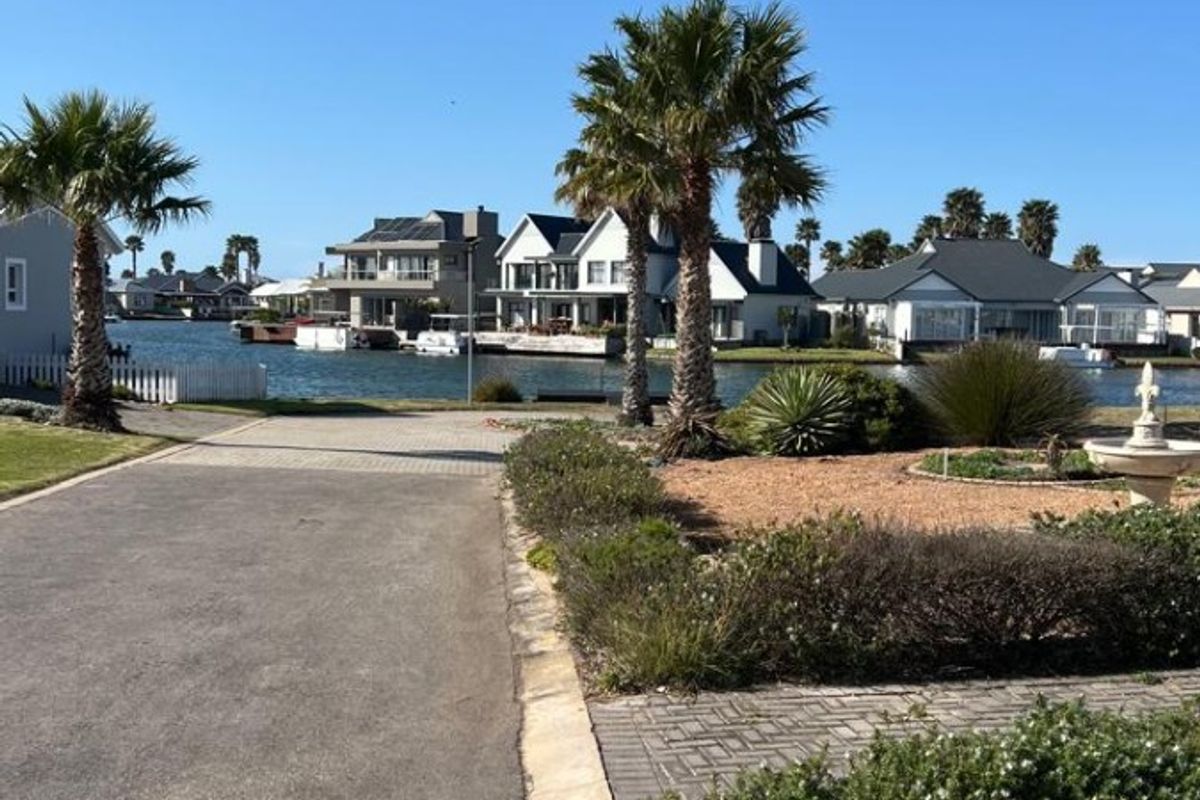 A scenic view of a residential waterfront area featuring a paved driveway, landscaped garden, and a decorative fountain overlooking the canal.