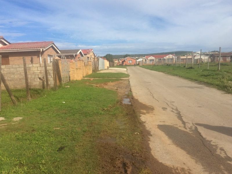 A street view showing the residential neighbourhood approach with asphalt road, brick boundary walls, and wire fencing along the property line.