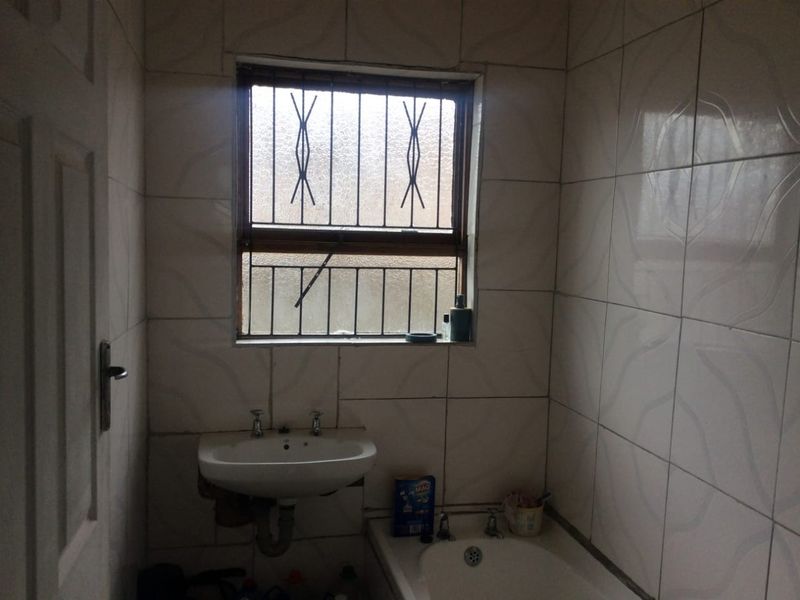 A dated bathroom featuring a wall-mounted basin, built-in bathtub, and full-height wall tiling, requiring cosmetic updates and maintenance to the plumbing and finishes.