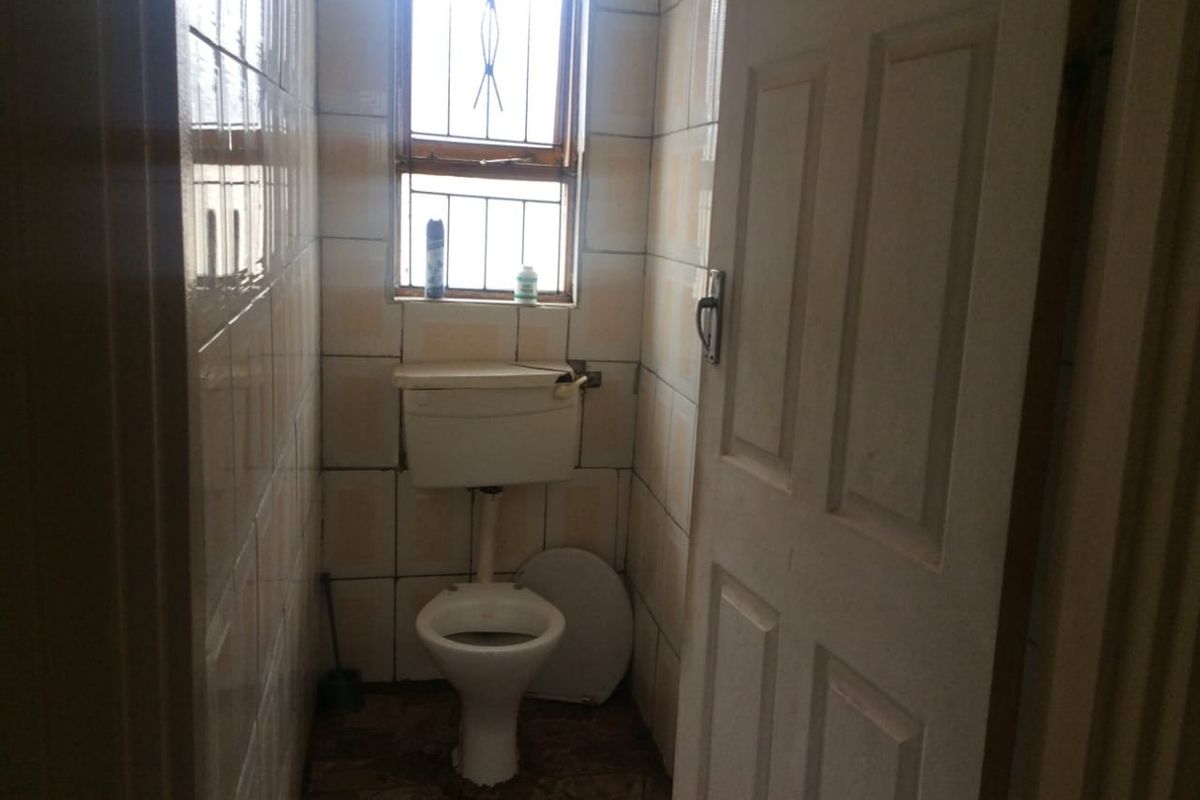 A small, dated bathroom featuring tiled walls and floor, equipped with a standard toilet and a window with security bars.
