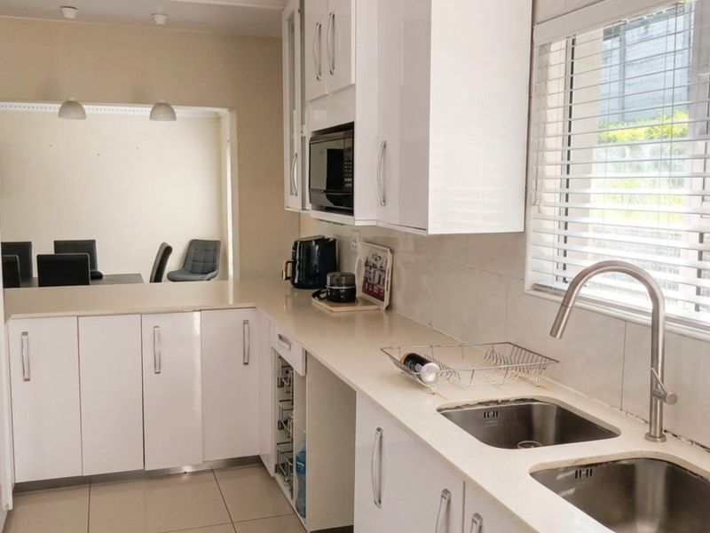 Modern kitchen featuring white gloss cabinetry, stone countertops, double stainless steel sink, and built-in microwave, with an open-plan flow to the dining area.