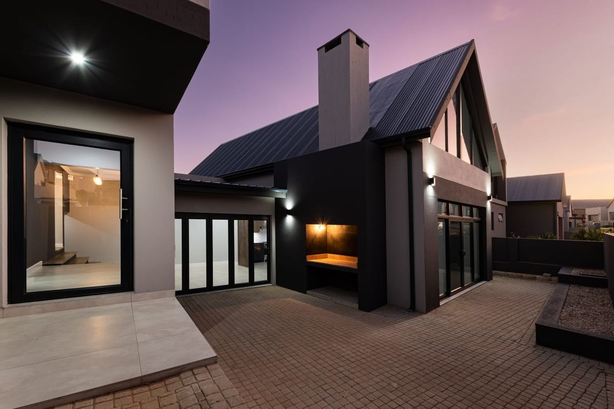 Modern rear exterior featuring a paved courtyard, built-in braai area, and contemporary architectural design with large glass doors and a striking metal roof.