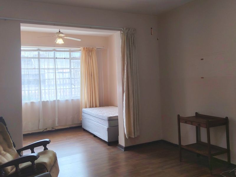A living area featuring laminate flooring, a ceiling fan, and a large window, currently showing signs of wear on the walls.