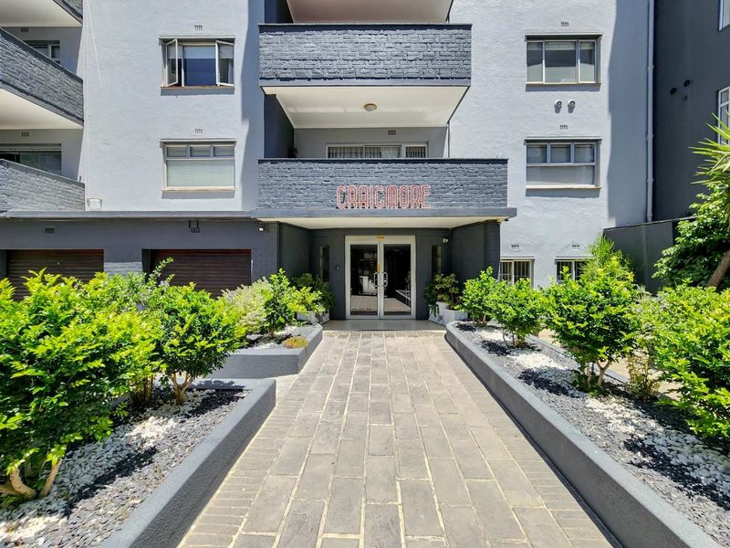 The front entrance of the Craigmore apartment building featuring a paved walkway, landscaped garden beds, and a well-maintained brick facade.