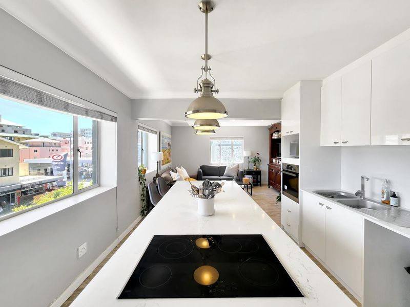 Modern open-plan kitchen and living area featuring a central island with electric cooktop, white cabinetry, and integrated appliances, overlooking an urban street view.