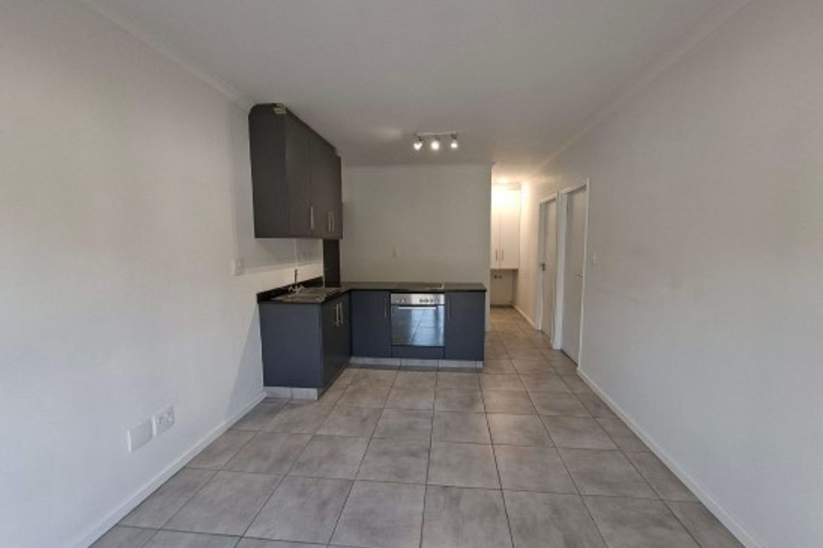 An open-plan kitchen area featuring modern dark cabinetry, integrated oven, and neutral tiled flooring in a clean, vacant apartment setting.