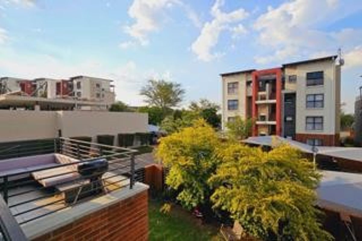 An elevated view of a residential complex featuring a private balcony with metal railings, overlooking a landscaped garden and covered parking area.
