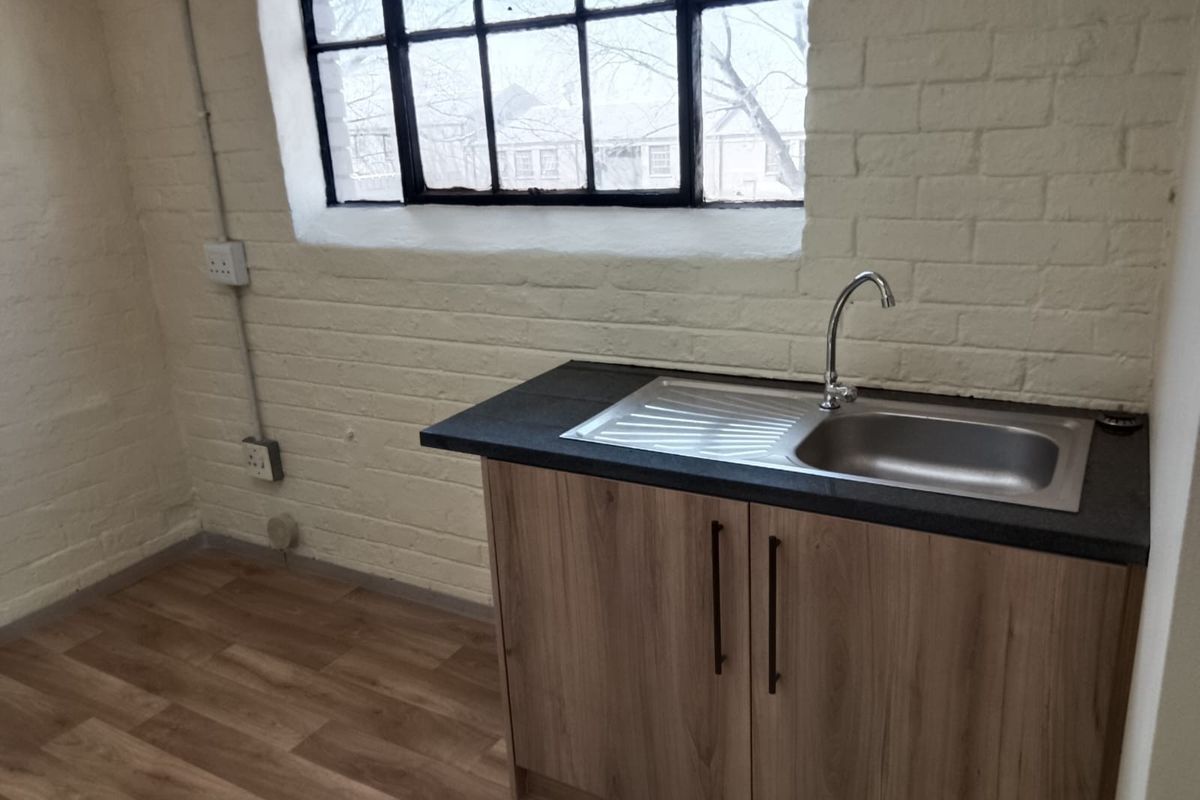 A compact industrial-style kitchenette featuring a stainless steel sink, wood-finish base cabinetry, and exposed painted brick walls with a large window.