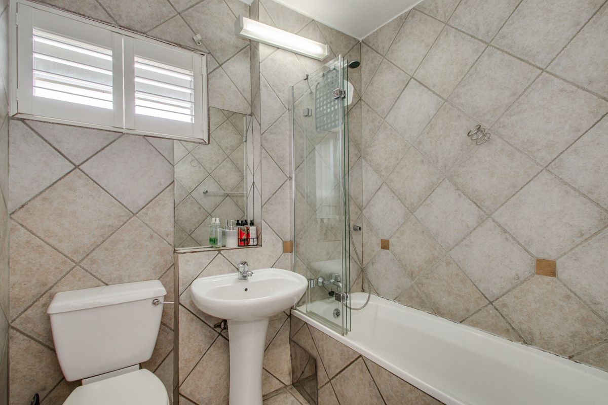A functional bathroom featuring a pedestal basin, bathtub with a glass shower screen, and tiled walls with a shutter window.