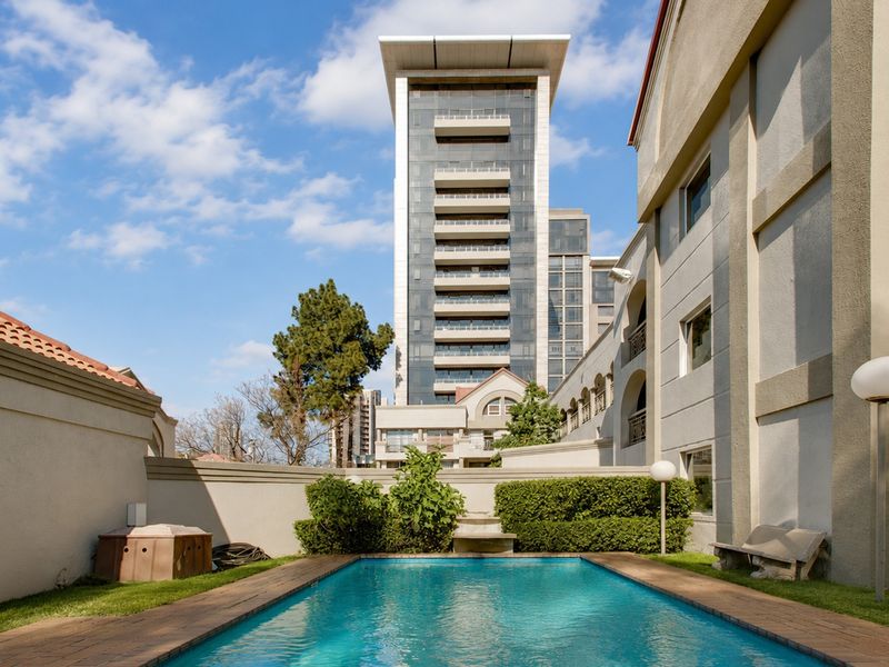 A well-maintained outdoor swimming pool area featuring brick coping, landscaped garden borders, and a high boundary wall, set against a modern apartment building.
