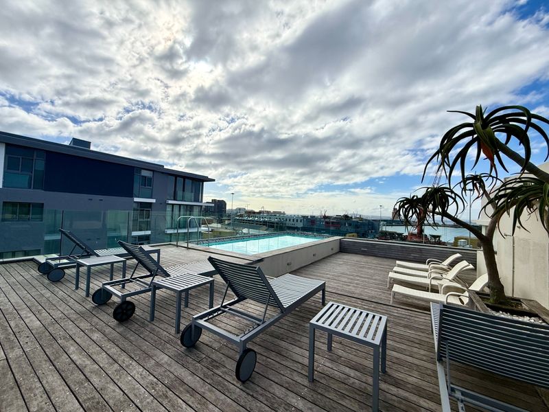Rooftop pool area featuring timber decking, glass balustrades, and expansive urban and harbour views.