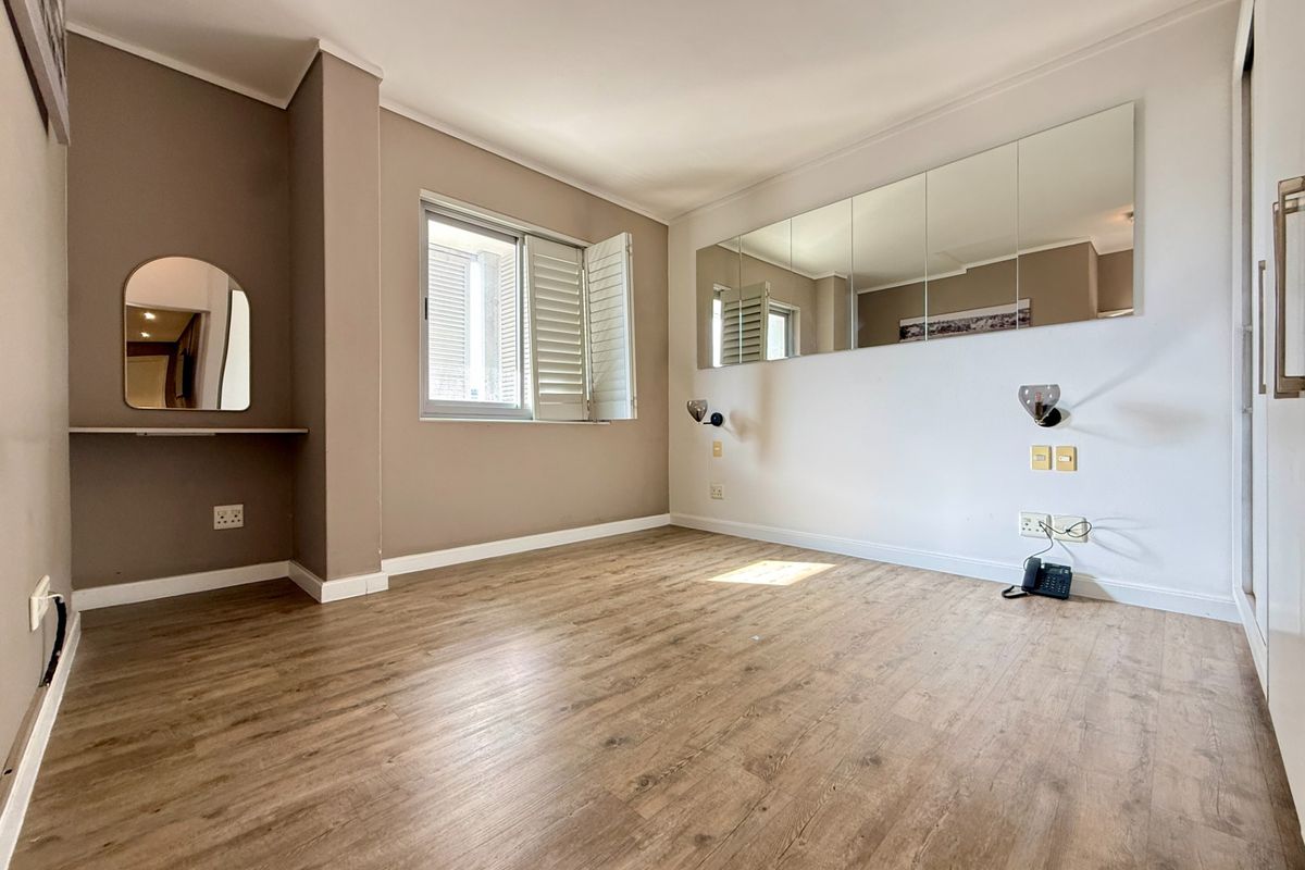A bright, empty bedroom featuring laminate flooring, built-in cupboards, window shutters, and wall-mounted vanity and bedside lighting fixtures.